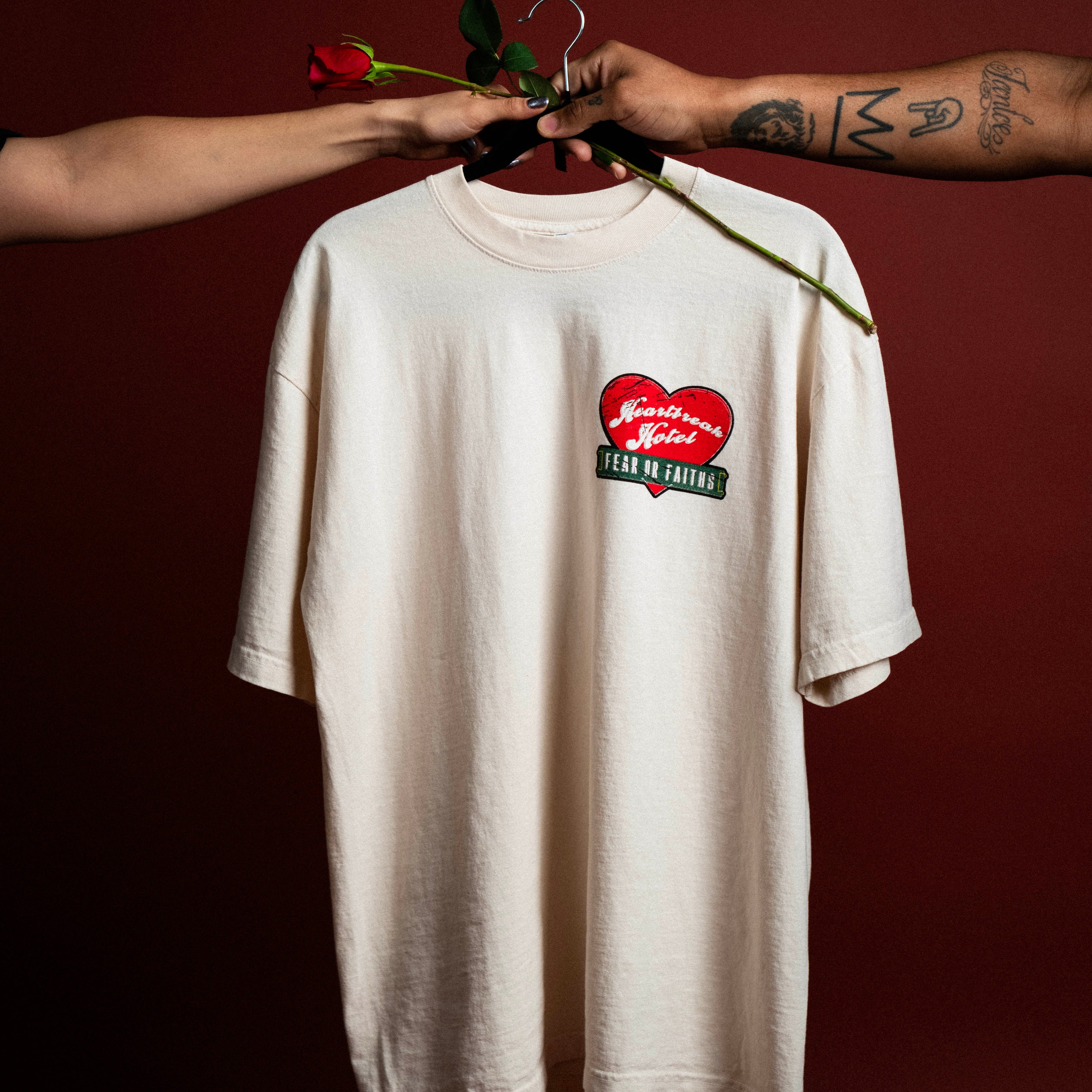 A white tee with a red heart being held up by two hands in front of a red background.